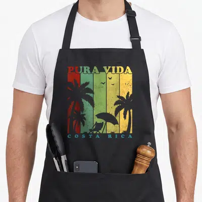 Costa summer aprons - Retro beach scene with PURA VIDA text, red green yellow stripes, palm trees, chair, umbrella — shown on a black cotton canvas apron against a white background