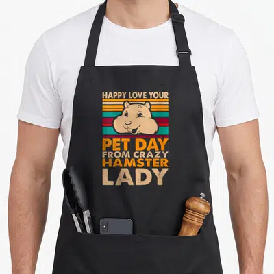 Cute hamster pet apron - Retro tan hamster head over orange teal and red stripes with stacked text reading HAPPY LOVE — shown on a black cotton canvas apron against a white background