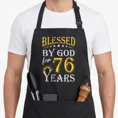 76th birthday apron - Yellow and white stacked text Blessed by God for 76 Years with five stars and sunburst — shown on a black cotton canvas apron against a white background
