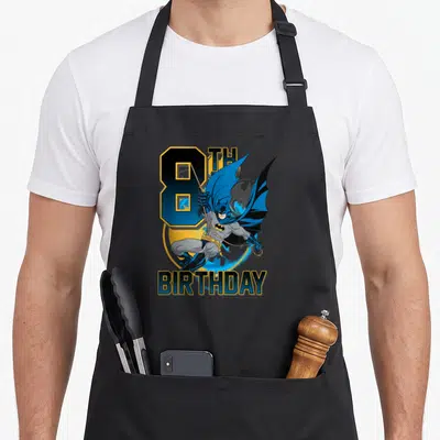8th bat birthday apron - Batman swinging over large blue number 8 and yellow circle with 8TH BIRTHDAY text — shown on a black cotton canvas apron against a white background