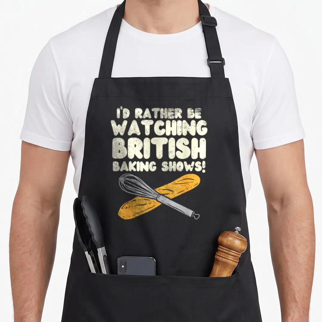 Show baker apron - I'D RATHER BE WATCHING BRITISH BAKING SHOWS text above crossed whisk and baguette in orange and grey — shown on a black cotton canvas apron against a white background