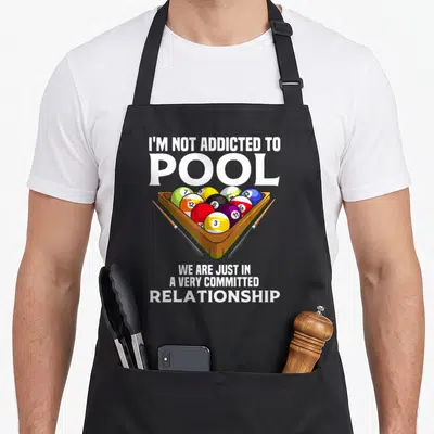 Addicted billiards apron - Cartoon pool balls in a brown rack with two cues and white commitment relationship text — shown on a black cotton canvas apron against a white background