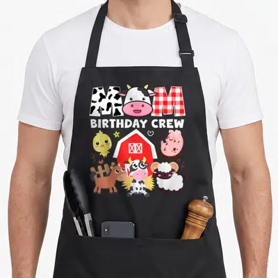 Crew cow print apron - MOM BIRTHDAY CREW text with cow head, red barn, and farm animals like pig and sheep — shown on a black cotton canvas apron against a white background