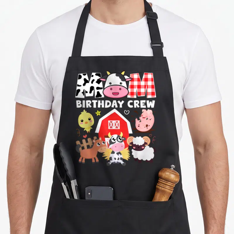 Crew cow print apron - MOM BIRTHDAY CREW text with cow head, red barn, and farm animals like pig and sheep — shown on a black cotton canvas apron against a white background