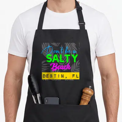 Salty florida apron - Stacked neon text reading Don't be a SALTY Beach with grey palm leaves and yellow destination bar — shown on a black cotton canvas apron against a white background