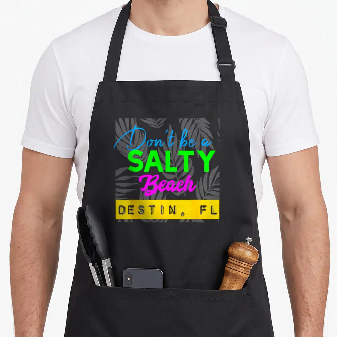 Salty florida apron - Stacked neon text reading Don't be a SALTY Beach with grey palm leaves and yellow destination bar — shown on a black cotton canvas apron against a white background