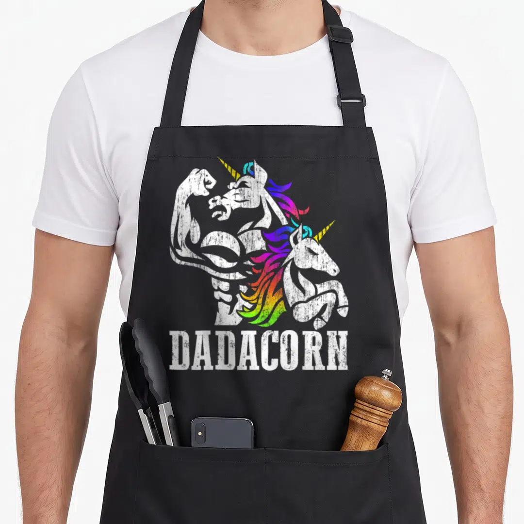 Dadacorn unicorn apron - Muscular unicorn flexing with smaller head and DADACORN text in distressed rainbow colors — shown on a black cotton canvas apron against a white background