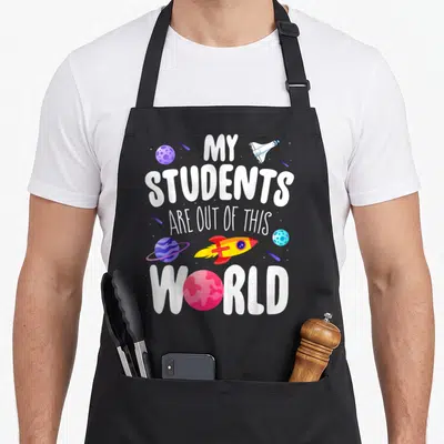 Student space teacher apron - My Students Are Out Of This World text with cartoon planets and rockets in purple blue — shown on a black cotton canvas apron against a white background