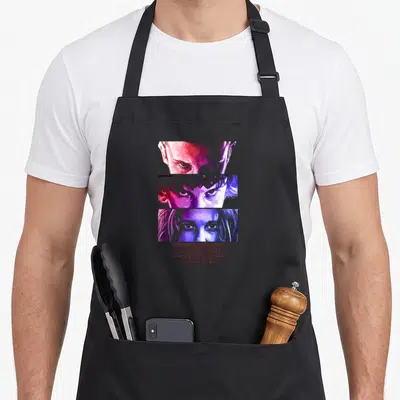 Stare novelty apron - Three stacked face strips in red, purple, and blue with centered Stranger Things text — shown on a black cotton canvas apron against a white background