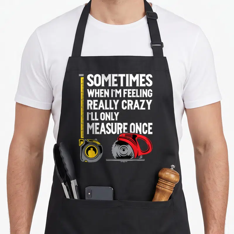 Ill woodworking apron - Stacked white text with yellow tape measure and red circular saw illustration — shown on a black cotton canvas apron against a white background