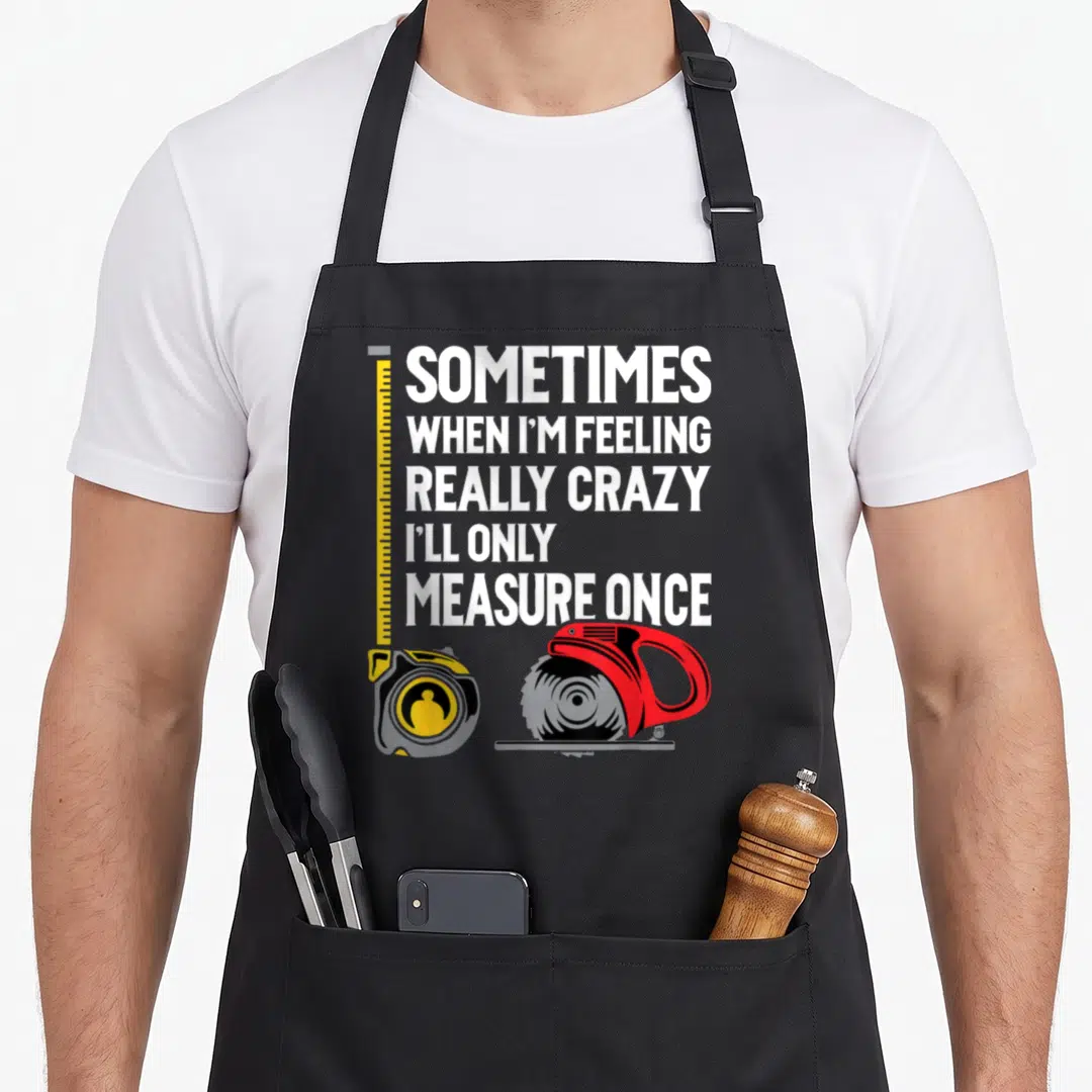 Ill woodworking apron - Stacked white text with yellow tape measure and red circular saw illustration — shown on a black cotton canvas apron against a white background