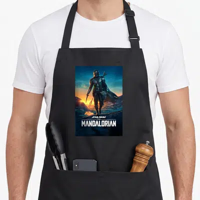 Season star war apron - Mandalorian figure holding Grogu in rocky sunset landscape with white text at bottom — shown on a black cotton canvas apron against a white background