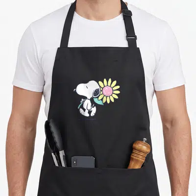 Baseball daisy apron - Walking cartoon Snoopy holding large pink and yellow flower with light blue stem — shown on a black cotton canvas apron against a white background