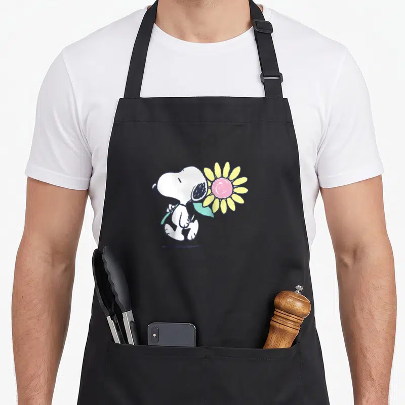 Baseball daisy apron - Walking cartoon Snoopy holding large pink and yellow flower with light blue stem — shown on a black cotton canvas apron against a white background