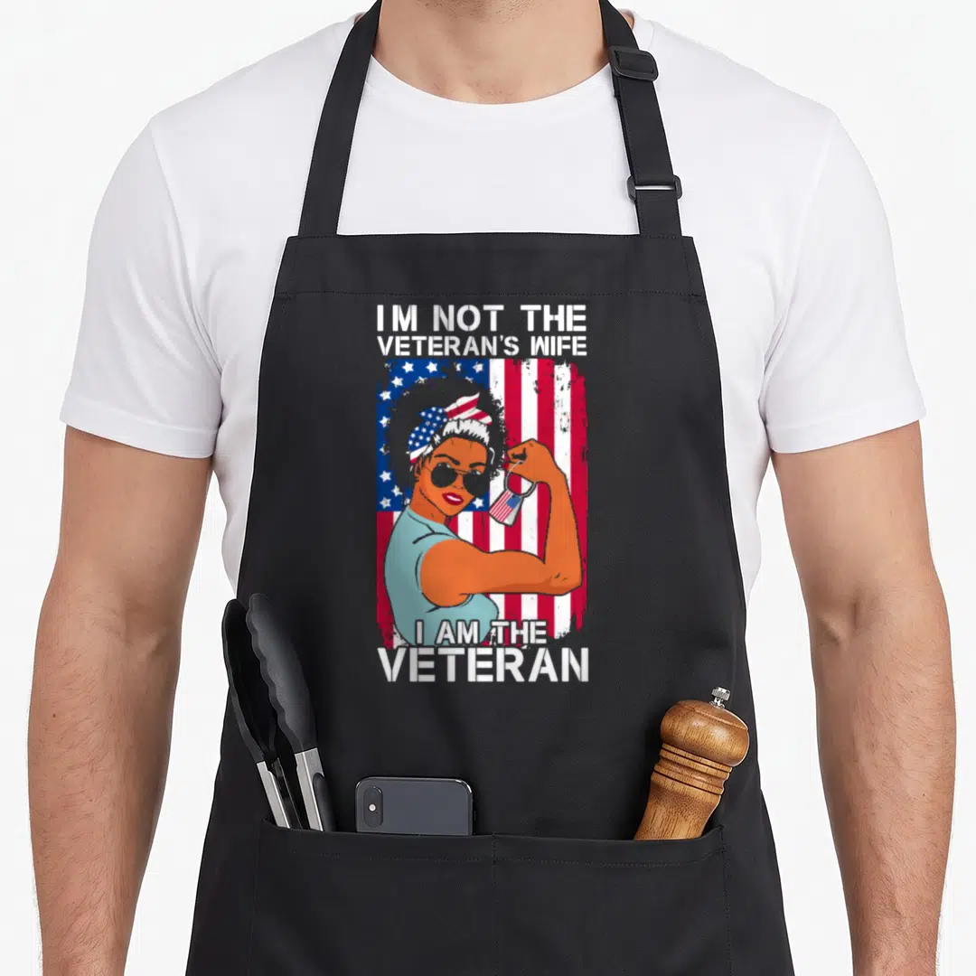 Veteran military patriotic apron - Woman flexing arm holding dog tag in front of distressed flag with text I am — shown on a black cotton canvas apron against a white background