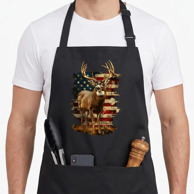 Deer 4th of july apron - Brown deer standing in front of distressed red white and blue American flag with grass at feet — shown on a black cotton canvas apron against a white background