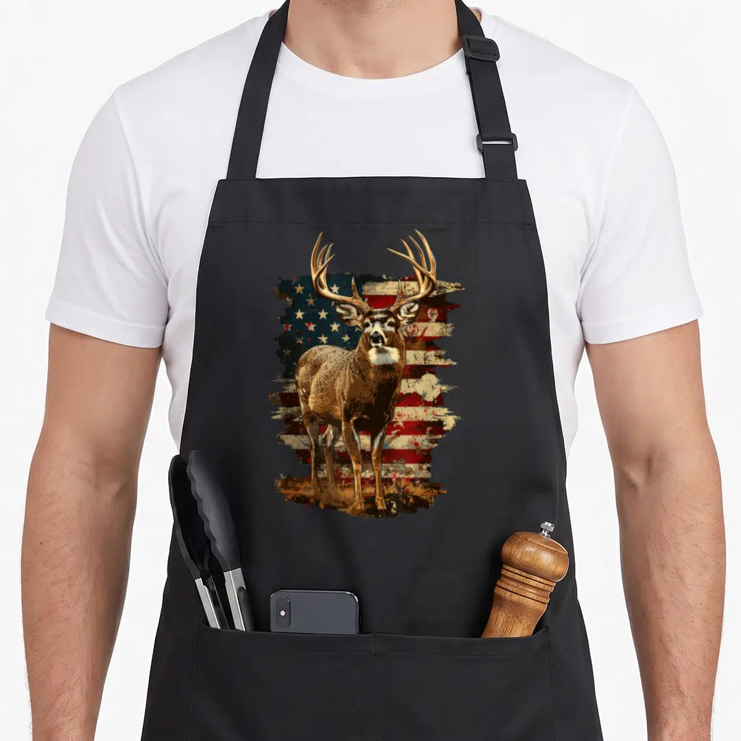 Deer 4th of july apron - Brown deer standing in front of distressed red white and blue American flag with grass at feet — shown on a black cotton canvas apron against a white background