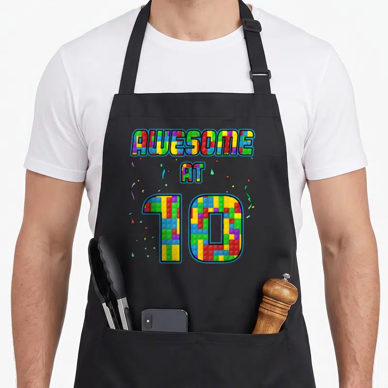10th birthday building aprons - AWESOME AT 10 text made of red blue yellow green and purple toy blocks with confetti — shown on a black cotton canvas apron against a white background