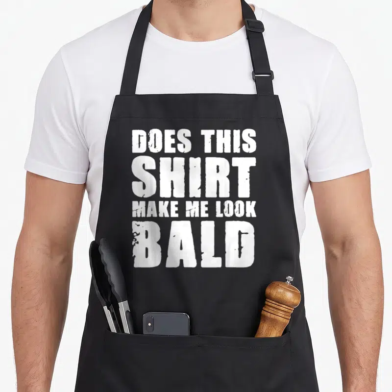 Doe husband funny apron - White stacked text reading DOES THIS SHIRT MAKE ME LOOK BALD on black fabric — shown on a black cotton canvas apron against a white background