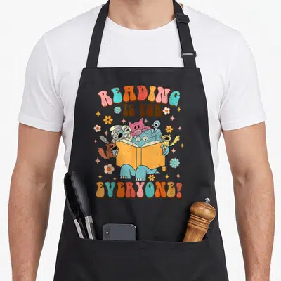 Bookish reading books apron - Colorful cartoon animals reading together under arched text surrounded by flowers — shown on a black cotton canvas apron against a white background