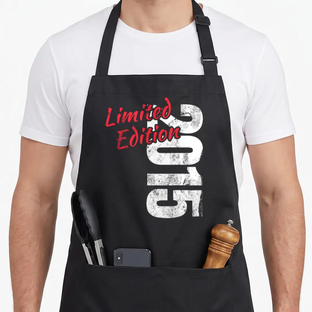 Edition anniversary birthday apron - Vertical white 2015 numbers with overlapping red Limited Edition script text — shown on a black cotton canvas apron against a white background