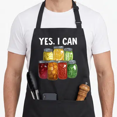 Season canning apron - YES, I CAN text above seven colorful cartoon canning jars — shown on a black cotton canvas apron against a white background