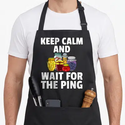 Pickling canning apron - KEEP CALM AND WAIT FOR THE PING text surrounding five colorful preserve jars in cartoon style — shown on a black cotton canvas apron against a white background