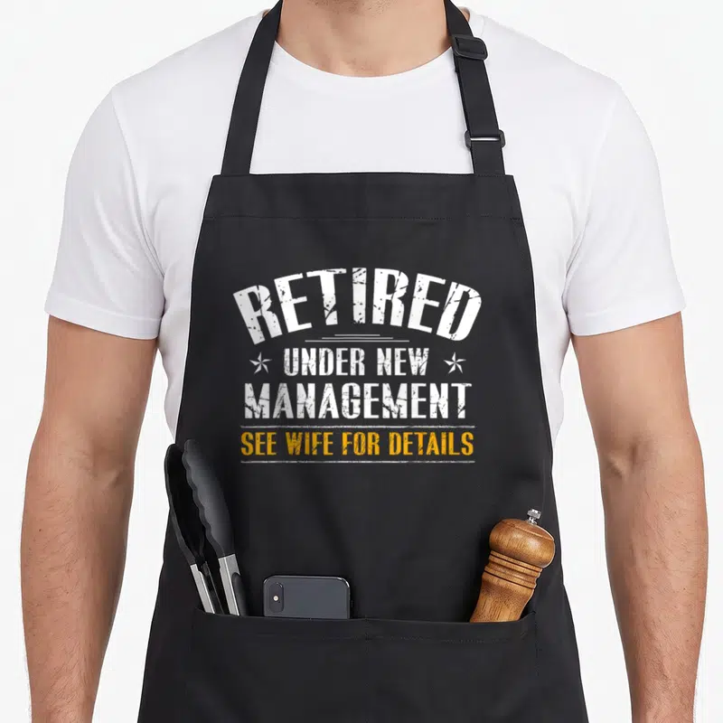 Retirement wife apron - White and yellow retro typography reading Retired Under New Management See Wife For Details — shown on a black cotton canvas apron against a white background