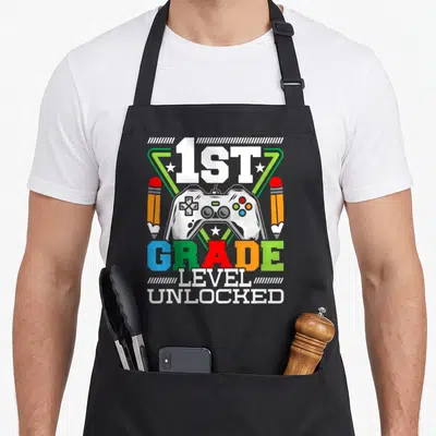 1st grade video game apron - Game controller flanked by two pencils with 1ST GRADE LEVEL UNLOCKED text in colorful — shown on a black cotton canvas apron against a white background
