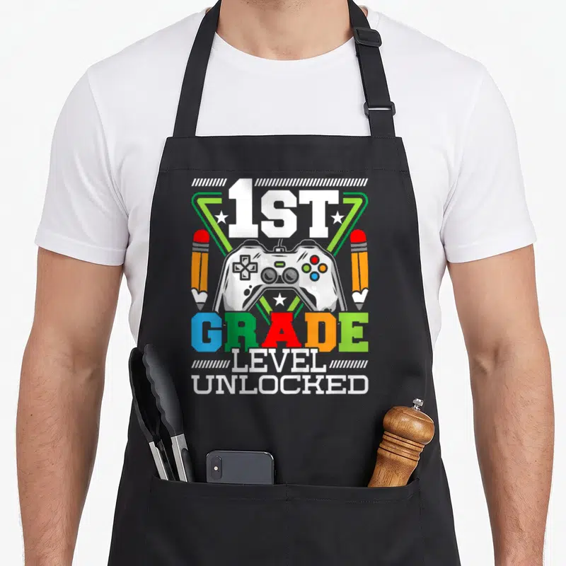 1st grade video game apron - Game controller flanked by two pencils with 1ST GRADE LEVEL UNLOCKED text in colorful — shown on a black cotton canvas apron against a white background