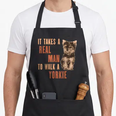 Walk yorkie apron - Stacked orange text beside sitting brown and tan cartoon Yorkshire Terrier illustration — shown on a black cotton canvas apron against a white background