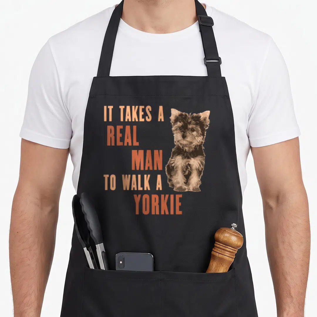 Walk yorkie apron - Stacked orange text beside sitting brown and tan cartoon Yorkshire Terrier illustration — shown on a black cotton canvas apron against a white background
