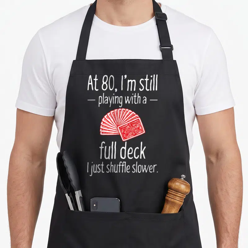 80th birthday apron - White text reading At 80 full deck with red fan of playing cards — shown on a black cotton canvas apron against a white background