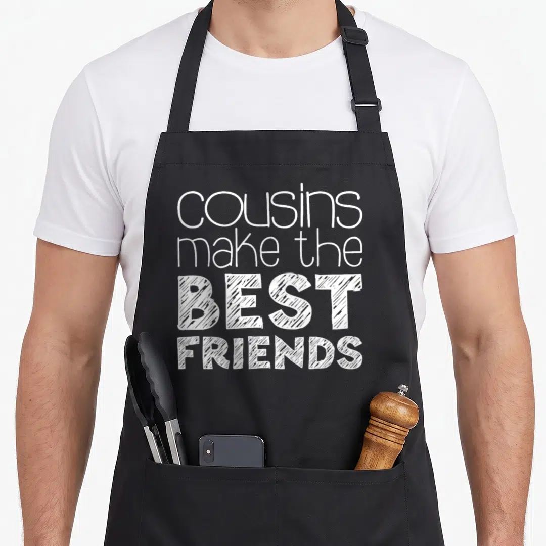 Cousin squad friends apron - White text reading cousins make the best friends stacked vertically on a dark background — shown on a black cotton canvas apron against a white background