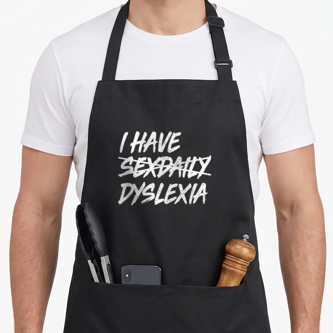 Sexdaily daily apron - White text I Have Dyslexia with sexdaily crossed out on black fabric — shown on a black cotton canvas apron against a white background