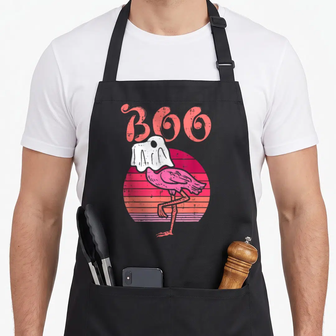 Flamingo birds apron - Coral BOO text above pink flamingo in white ghost sheet against striped pink and orange sun — shown on a black cotton canvas apron against a white background