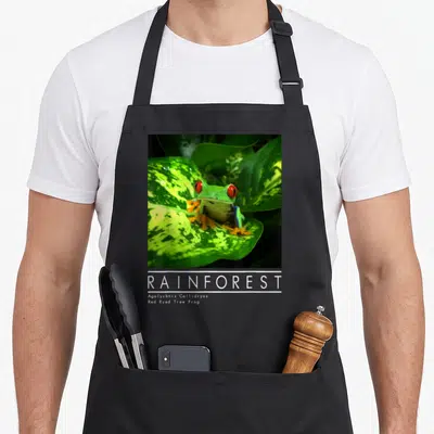 Amphibian tree apron - Square photo of green frog with red eyes on variegated leaf above white Rainforest text — shown on a black cotton canvas apron against a white background