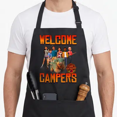 Jurassic happy camper apron - WELCOME and CAMPERS text framing people and a green dinosaur with Camp Cretaceous logo — shown on a black cotton canvas apron against a white background