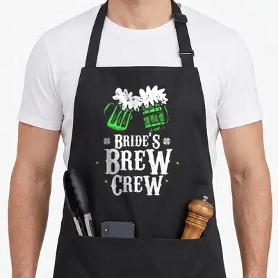 Bride st patrick day aprons - Two green beer mugs clink above stacked white text reading Bride's Brew Crew with small — shown on a black cotton canvas apron against a white background