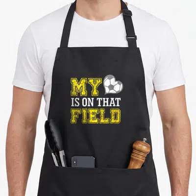 Field soccer mother's day aprons - Stacked yellow and white text reading MY IS ON THAT FIELD next to a white — shown on a black cotton canvas apron against a white background