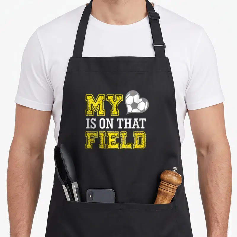 Field soccer mother's day aprons - Stacked yellow and white text reading MY IS ON THAT FIELD next to a white — shown on a black cotton canvas apron against a white background