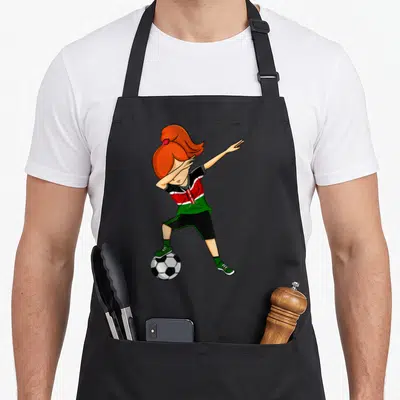 Dabbing kenyan boy apron - Cartoon girl in red green and black kit dabbing on soccer ball — shown on a black cotton canvas apron against a white background