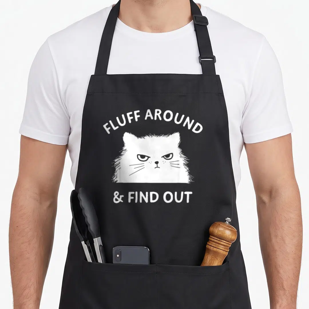 Fluff cat lady apron - White cartoon cat head with arched text reading FLUFF AROUND above and & FIND OUT below — shown on a black cotton canvas apron against a white background