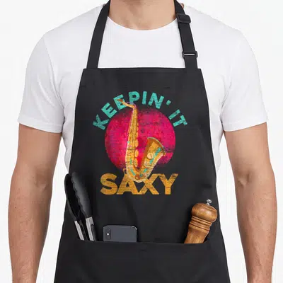 Sch slides apron - Retro saxophone over pink circle with teal and orange text reading KEEPIN' IT SAXY — shown on a black cotton canvas apron against a white background