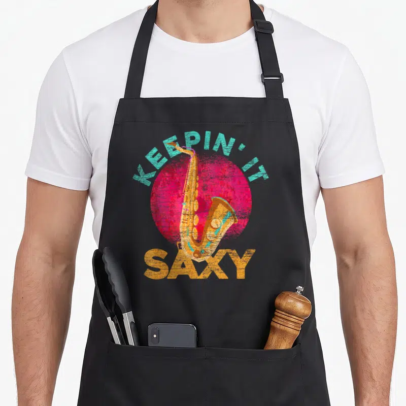 Sch slides apron - Retro saxophone over pink circle with teal and orange text reading KEEPIN' IT SAXY — shown on a black cotton canvas apron against a white background