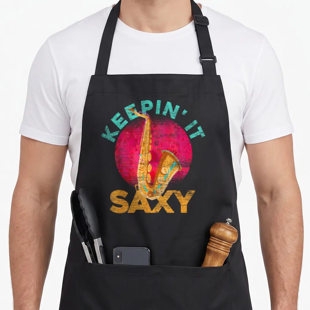 Sch slides apron - Retro saxophone over pink circle with teal and orange text reading KEEPIN' IT SAXY — shown on a black cotton canvas apron against a white background
