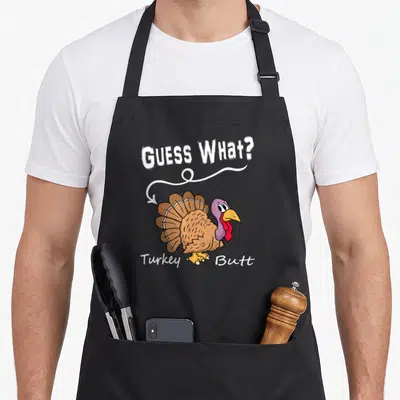 Guess thanksgiving apron - Cartoon brown turkey facing back with GUESS WHAT? and Turkey Butt text and white arrow — shown on a black cotton canvas apron against a white background