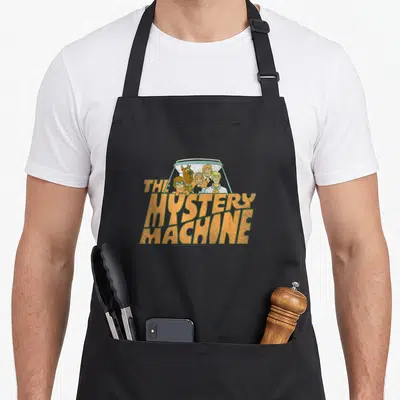 Mystery machine apron - Cartoon crew in van window above stacked orange text THE MYSTERY MACHINE — shown on a black cotton canvas apron against a white background