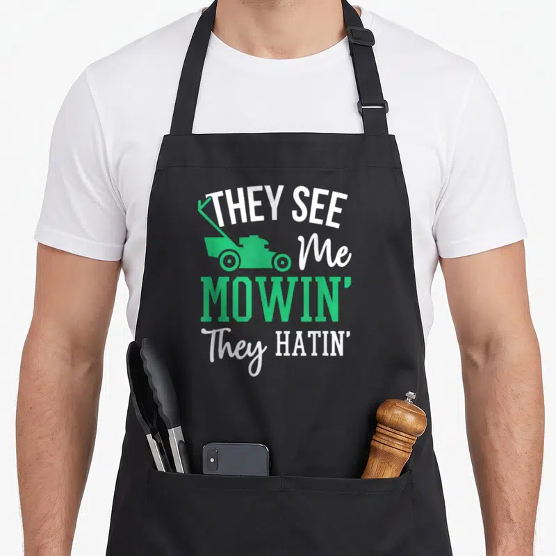 Hatin see me aprons - Stacked THEY SEE Me MOWIN They HATIN text with green push mower silhouette — shown on a black cotton canvas apron against a white background