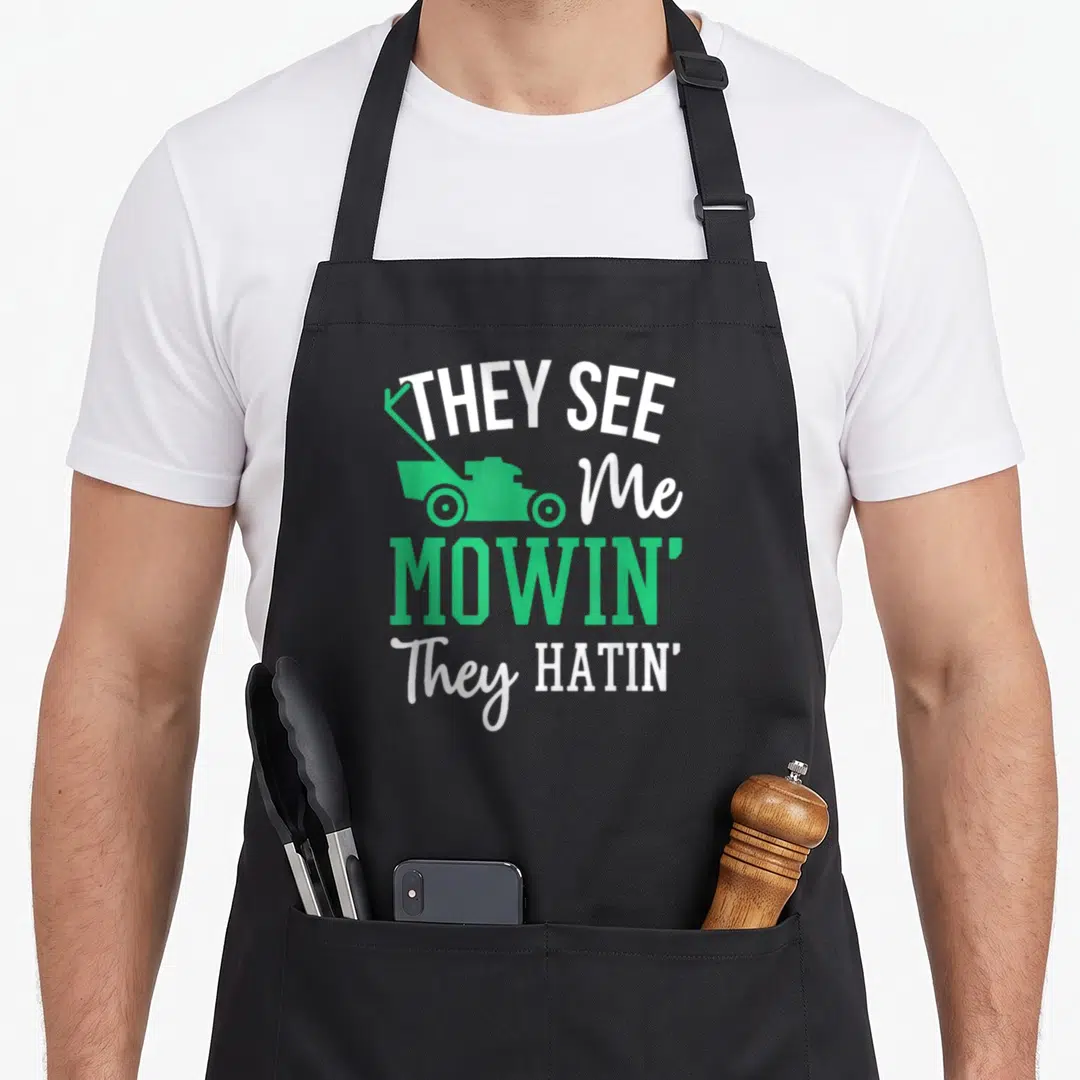 Hatin see me aprons - Stacked THEY SEE Me MOWIN They HATIN text with green push mower silhouette — shown on a black cotton canvas apron against a white background